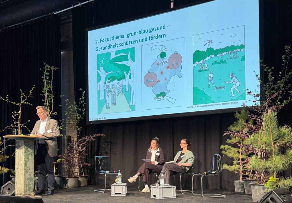 Auf einer Bühne spricht ein Mann an einem Rednerpult, während zwei Personen sitzen. Im Hintergrund wird eine Folie zum Fokusthema „grün-blau gesund“ mit Illustrationen zu Gesundheit und Stadtnatur gezeigt.