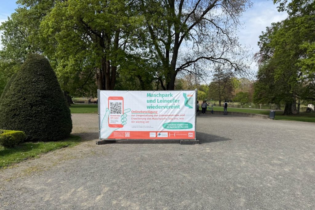 Ein großes Informationsbanner im Maschpark weist auf die Onlinebeteiligung zur Umgestaltung der Culemannstraße und Erweiterung des Maschparks hin. Das Banner steht auf einem breiten Kiesweg zwischen Bäumen und Grünflächen.