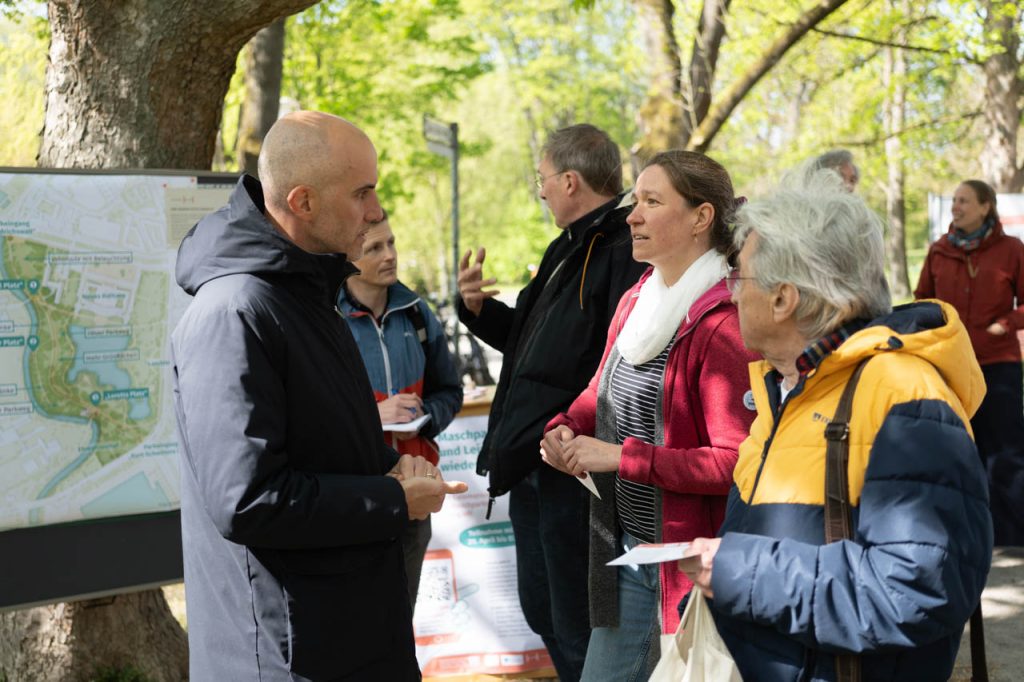Mehrere Personen stehen im Maschpark an einem Informationsstand mit Karte und Plakat. Sie sprechen miteinander über die geplante Umgestaltung und halten Informationsmaterial in den Händen.